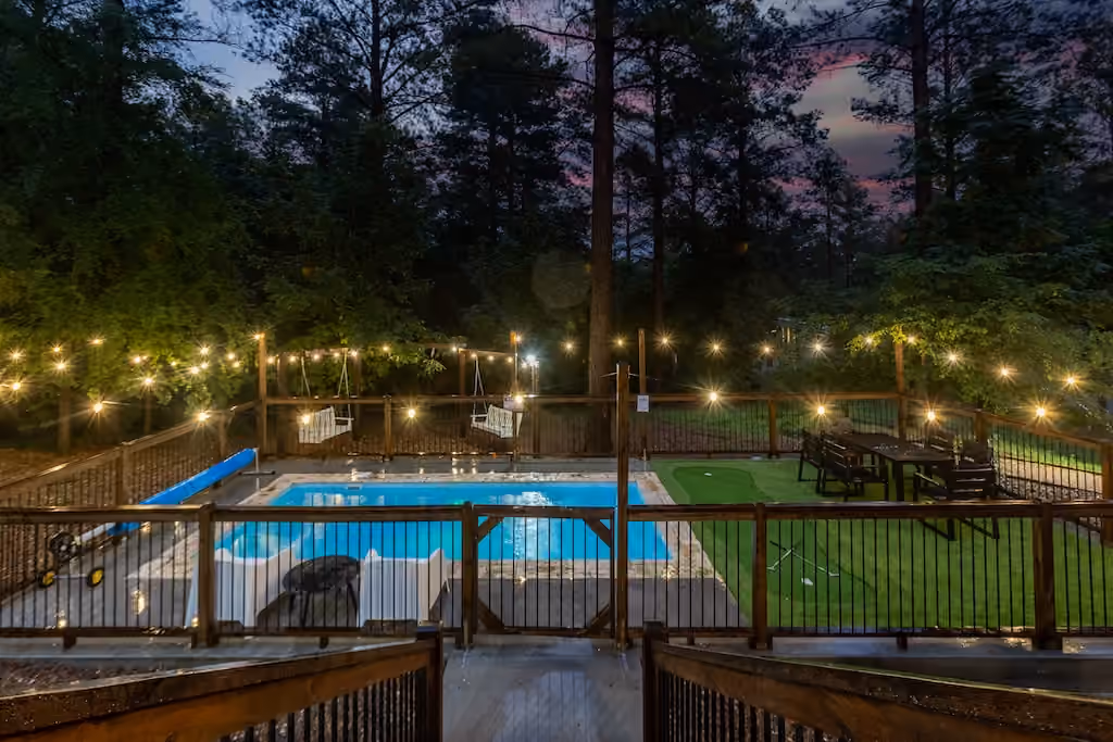 A backyard swimming pool and deck illuminated by string lights at dusk