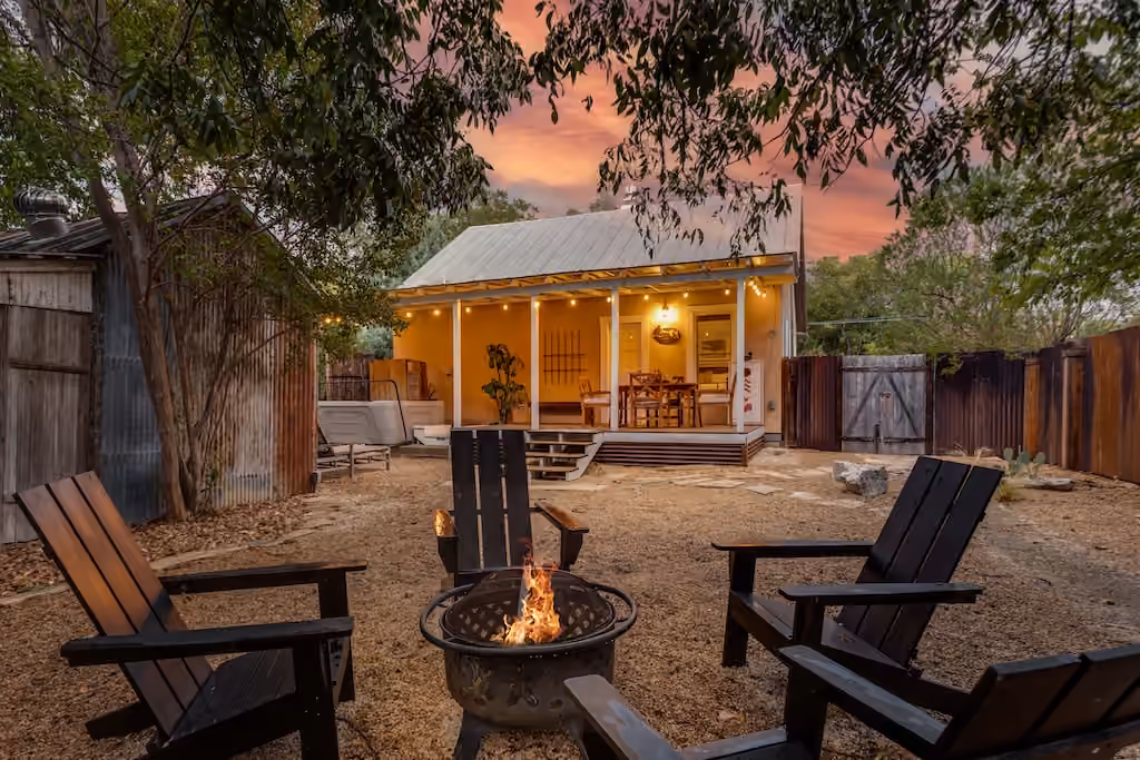 A backyard fire pit surrounded by chairs beneath trees at sunset